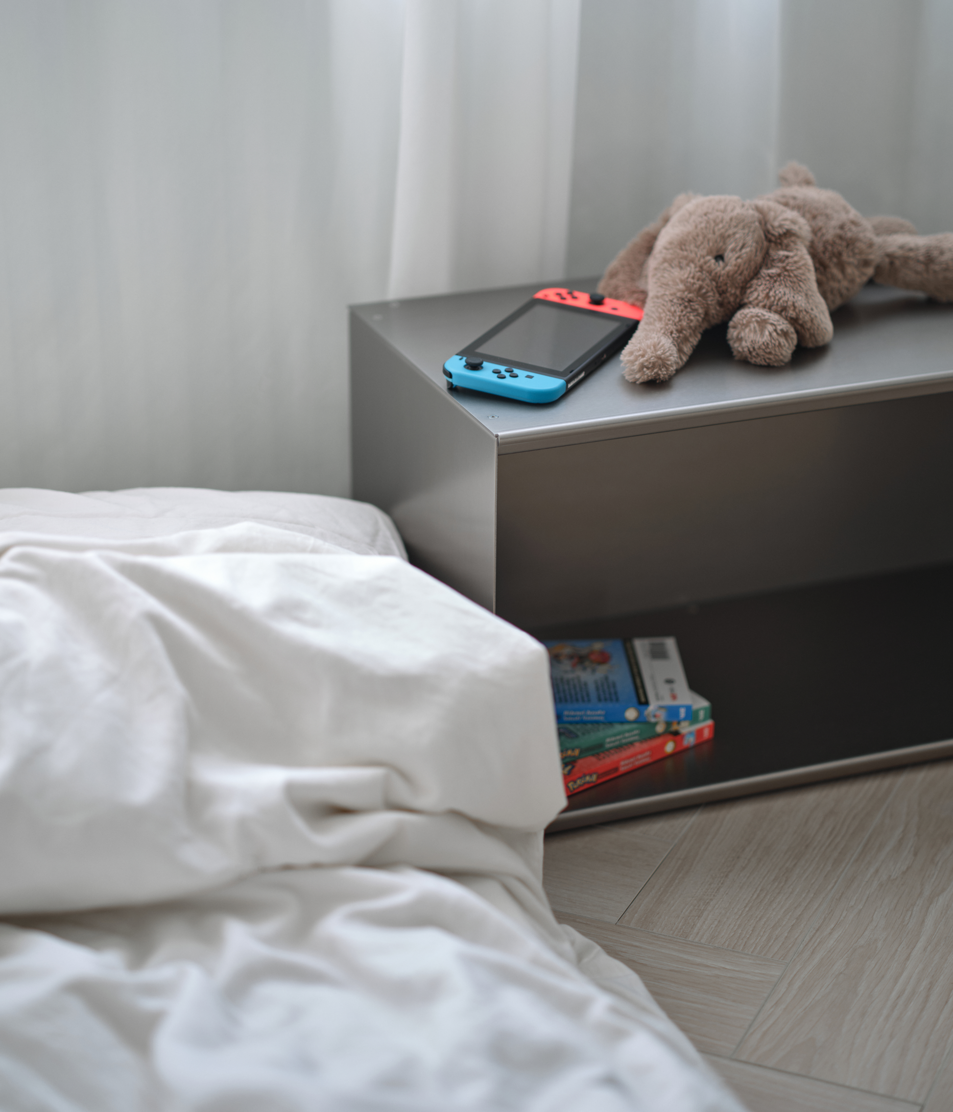 Small M85 aluminum bedside table with a teddy bear and gaming console next to a bed