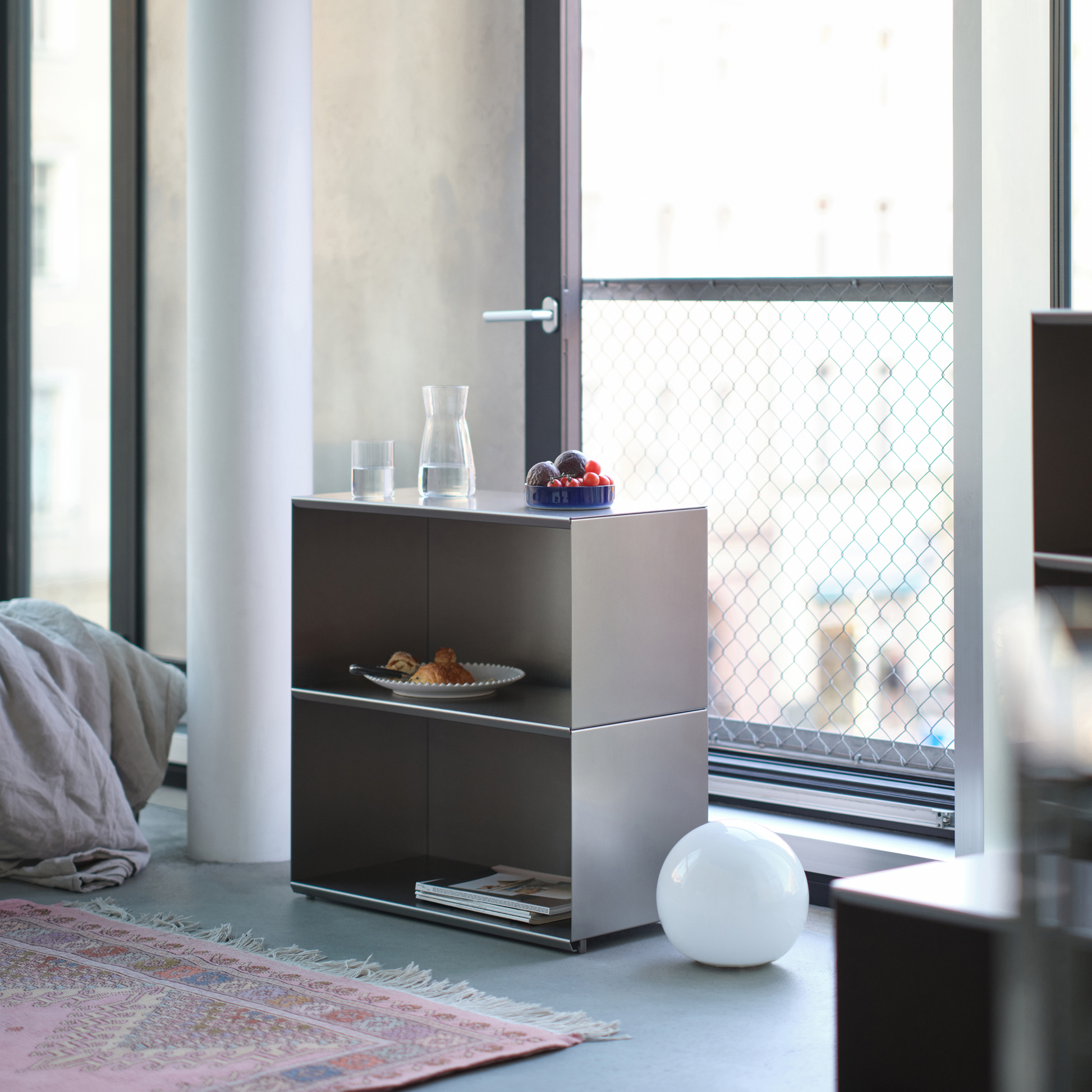 M85 modular aluminum side table with a glass of water and a plate of food in a living room with large windows.